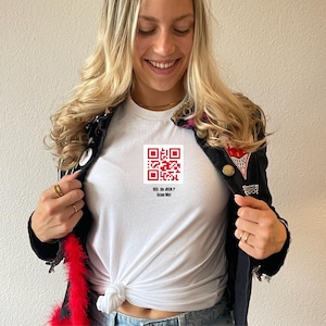 Könnte beinhalten: Weißes T-Shirt mit einem rot-weißen QR-Code-Grafik. Der Text unter dem QR-Code lautet "HIS OR JEDI? Scan Me!". Das Shirt wird mit einer schwarzen Jacke mit Aufnähern und einer roten Federboa getragen.