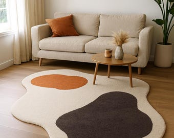 Op de afbeelding: Een crèmekleurig, abstract gevormd vloerkleed met twee grote, onregelmatige vlekken in bruin en oranje. Het vloerkleed ligt op een houten vloer in een woonkamer, met een bank, salontafel en kunstwerk op de achtergrond.