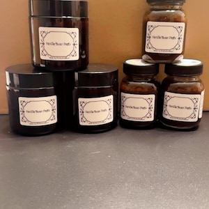 May include: An assortment of small glass jars with black lids. Each jar has a white label with the text "Vanilla Bean Perfume" in a decorative font. The jars are filled with a dark brown liquid. The jars vary in size and are arranged on a gray surface.