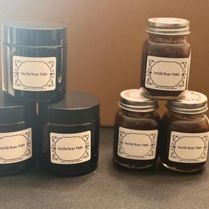 May include: An assortment of jars filled with vanilla bean paste. The jars are black with silver lids and white labels with decorative borders. The labels read "Vanilla Bean Paste". The jars are arranged on a dark surface.