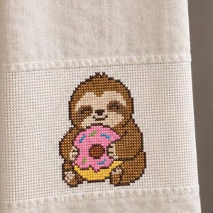以下が含まれることがあります： カラフルなスプリンクルがのったピンク色のドーナツを持つ、茶色のナマケモノのクロスステッチ刺繍が施されたベージュのキッチンタオル。タオルには質感のある布地バンドがあり、その中央にナマケモノのデザインが配置されています。