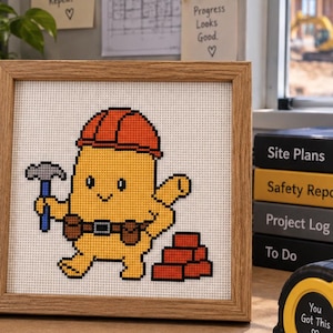 May include: A square wooden frame containing a cross-stitch pattern of a yellow cartoon character wearing an orange hard hat, holding a blue hammer, and standing next to a stack of red bricks, all set against a white fabric background.