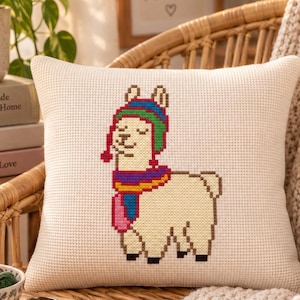 May include: A square off-white throw pillow featuring a cross-stitch design of a cream-colored llama wearing a multi-colored striped winter hat and a matching scarf. The llama is depicted in a pixelated style with brown hooves and ears.