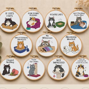 May include: Ten circular embroidery hoops featuring cross-stitch designs of cats with humorous text. Each hoop displays a unique cat illustration with captions like IF I FITS I SITS, BOW DOWN PEASANT, YOU'RE LATE, and NO WITNESSES on white fabric.