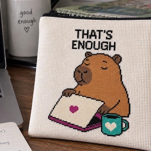 May include: A square, off-white zippered pouch featuring a cross-stitch design of a brown capybara sitting at a laptop with a pink heart. The text THAT'S ENOUGH is printed above the capybara, and a teal mug with a white heart sits beside it.