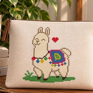 May include: A rectangular off-white fabric pouch featuring a cross-stitch design of a cream-colored llama with a colorful saddle, a multi-colored pom-pom necklace, and a small red heart above its head, standing on a patch of green grass.