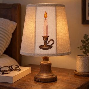 May include: A table lamp with a textured, brown, pedestal-style base and a tapered, light-colored fabric shade. The shade features a centered cross-stitch embroidery design of a lit candle in a holder with a yellow and orange flame.