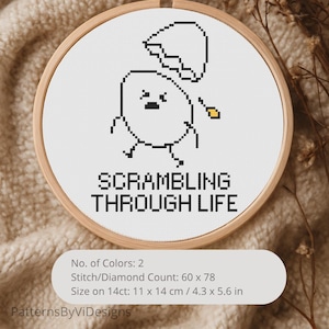 May include: A cross-stitch embroidery in a wooden hoop features a pixelated egg character with the text "SCRAMBLING THROUGH LIFE." The design includes a cracked eggshell above the egg. The finished size is 4.3 x 5.6 inches.