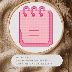 Puede incluir: Un diseño de punto de cruz rosa de un bloc de notas en un bastidor de bordado de madera. El bloc de notas tiene tres líneas horizontales y una encuadernación en espiral. El diseño mide 13 x 7 cm. El texto de la imagen dice "PatternsByViDesigns".
