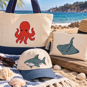 May include: A beige canvas tote bag with a red octopus graphic, a beige baseball cap with a blue shark graphic, and a beige zippered pouch with a blue stingray graphic. The pouch features a small starfish charm and a blue and beige tassel.