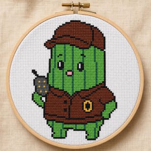 May include: A cross-stitch embroidery in a round wooden hoop featuring a green cactus character wearing a brown cap and a matching brown jacket. The cactus has a small pink mouth, black eyes, and is holding a dark gray walkie-talkie device.