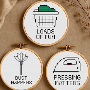 Könnte beinhalten: Drei runde Stickrahmen mit Kreuzstichmustern. Einer zeigt einen Wäschekorb mit dem Text "LOADS OF FUN". Ein anderer zeigt einen Staubwedel mit "DUST HAPPENS". Der letzte zeigt ein Bügeleisen mit "PRESSING MATTERS".
