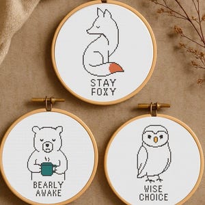 May include: Three circular embroidery hoops with cross-stitch designs. The top hoop shows a fox with the text "Stay Foxy." The middle hoop depicts a bear with a mug and the text "Bearly Awake." The bottom hoop displays an owl with the text "Wise Choice."