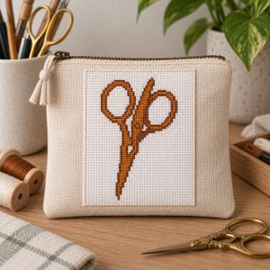 May include: A beige canvas zippered pouch featuring a centered white square panel with a brown cross-stitch embroidery design of a pair of scissors. The pouch has a matching beige fabric zipper pull with a small tassel at the end of the metal zipper.
