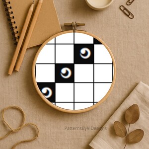May include: An embroidery hoop featuring a black and white checkered pattern with three curved, eye-like designs. The hoop is set against a neutral background with a notebook, pencils, a candle, and other craft supplies.