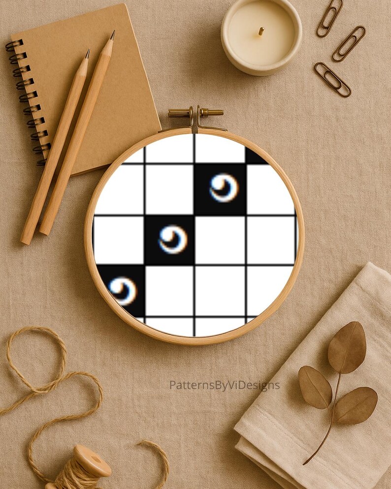 May include: An embroidery hoop featuring a black and white checkered pattern with three curved, eye-like designs. The hoop is set against a neutral background with a notebook, pencils, a candle, and other craft supplies.