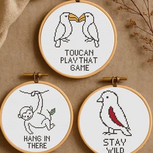 May include: Three circular embroidery hoops with cross-stitch designs. One shows two toucans and the text "TOUCAN PLAY THAT GAME." Another depicts a monkey with the text "HANG IN THERE." The last shows a bird with the text "STAY WILD."