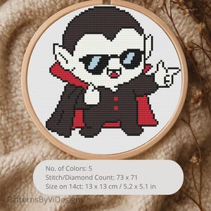 Peut inclure: Une broderie au point de croix dans un cercle en bois représente un vampire de dessin animé portant des lunettes de soleil et une cape, pointant du doigt. Le motif utilise du noir, du rouge, du blanc et des couleurs chair. La taille est de 13 x 13 cm. Le texte "PatternsByViDesigns" est en bas.