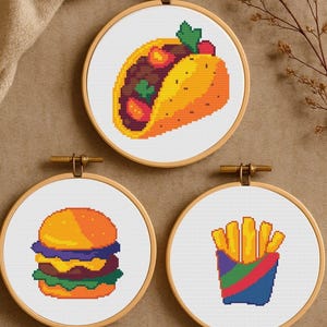 Peut inclure: Trois cercles de broderie au point de croix représentant des aliments. Le cercle supérieur affiche un taco avec des détails jaunes, oranges et bruns. En dessous, un burger avec des éléments orange, violets, verts et jaunes. Le dernier cercle montre un cornet de frites.