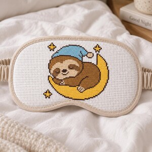 Puede incluir: Un antifaz para dormir blanco con un diseño de punto de cruz de un perezoso marrón con un gorro de dormir azul, descansando sobre una luna creciente amarilla rodeada de pequeñas estrellas amarillas. El antifaz tiene un borde beige y elástico.