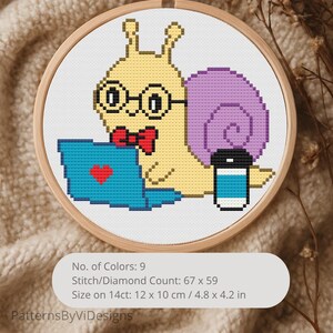 May include: Cross-stitch embroidery in a wooden hoop features a cartoon snail wearing glasses and a bow tie, working on a blue laptop with a heart. The snail has a purple shell and is next to a coffee cup. The design is 4.8 x 4.2 inches.