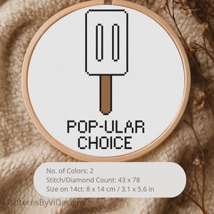 May include: Cross-stitch embroidery in a wooden hoop features a popsicle design with the text "POP-ULAR CHOICE." The popsicle is white with a black outline and a brown stick. The design measures 3.1 x 5.6 inches.
