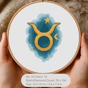 May include: A cross-stitch embroidery of the Taurus zodiac symbol in a wooden hoop. The design features a gold Taurus symbol on a blue and teal background with stars and a crescent moon. The size is 14 x 19 cm.