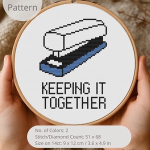 May include: A cross-stitch pattern in a wooden hoop features a pixelated stapler design in black, white, and blue. The text "KEEPING IT TOGETHER" is below the stapler. The pattern size is 3.6 x 4.9 inches.