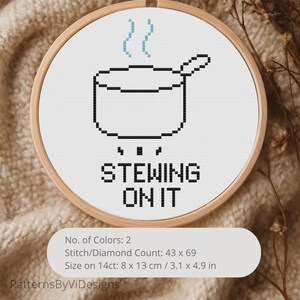 Peut inclure: Une broderie au point de croix dans un cercle en bois représente une marmite noire et blanche avec de la vapeur et le texte "STEWING ON IT". Le motif mesure 8 x 13 cm et utilise deux couleurs. Le fond est un tissu beige texturé.
