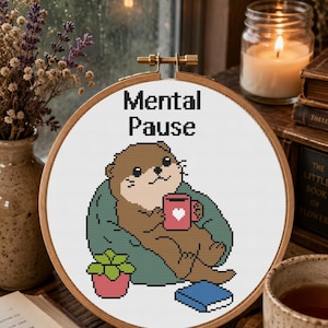 Könnte beinhalten: Ein runder Holzstickrahmen mit einem Kreuzstichmuster eines braunen Otters, der in einem grünen Sessel sitzt und eine rosa Tasse mit einem weißen Herz hält. Der Text Mental Pause ist darüber gestickt, ergänzt durch eine kleine Pflanze und ein blaues Buch.