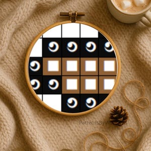 May include: A cross-stitch embroidery hoop with a geometric pattern in black, white, and brown. The design features squares and eye-like shapes. The hoop is set on a beige knitted surface, with a cup of coffee and a pinecone nearby.