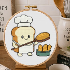 Può includere: Un telaio da ricamo circolare in legno contenente un motivo a punto croce di una fetta di pane antropomorfa sorridente che indossa un cappello da chef e un grembiule bianchi. Il personaggio tiene una pala con un pane, accanto a una ciotola verde di panini.