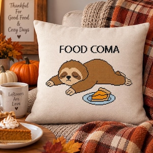 Puede incluir: Un cojín cuadrado de color beige con un gráfico estilo punto de cruz de un perezoso marrón tumbado, con el texto FOOD COMA impreso encima y un trozo de tarta naranja en un plato azul situado delante del perezoso en la funda del cojín.