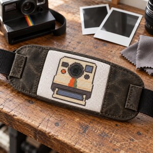 May include: A camera strap featuring a central white fabric panel with a cross-stitched pixel art design of an instant camera. The camera motif includes a black lens, a blue viewfinder, and a vertical stripe in red, orange, and green. The strap ends are made of dark brown, weathered-look material with black nylon webbing.