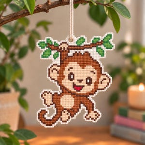May include: A cross-stitch ornament featuring a brown monkey with a cream-coloured belly hanging from a green leafy branch. The monkey has a smiling face with a red mouth and is suspended by a white string, showcasing a detailed pixelated design.
