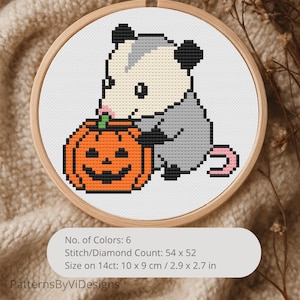 May include: Cross-stitch embroidery in a wooden hoop featuring a cartoon opossum with a gray body, black ears, and a pink tail, holding an orange jack-o'-lantern. The design is on white fabric. The text on the image says "PatternsByViDesigns".