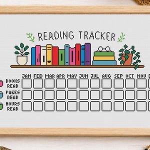 May include: A framed cross-stitch reading tracker with the words "Reading Tracker" at the top. The design includes a shelf with colorful books and potted plants. Below are boxes to track books, pages, and hours read.