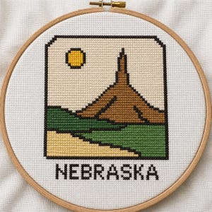 May include: A cross-stitch embroidery in a circular wooden hoop featuring a landscape with a brown rock formation, a yellow sun, green fields, and a tan foreground. The word NEBRASKA is stitched in black capital letters below the framed image.