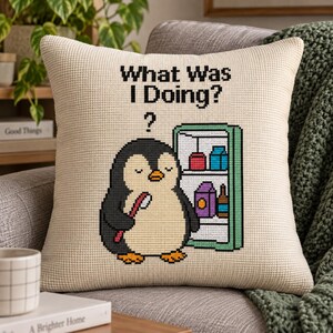 Può includere: Un cuscino quadrato color crema con un disegno a punto croce di un pinguino davanti a un frigorifero aperto. Il testo "What Was I Doing?" è stampato sopra il pinguino, che tiene uno spazzolino rosso sotto l'ala.