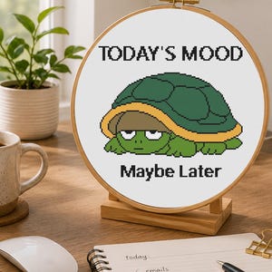 May include: A circular wooden embroidery hoop containing white fabric with a cross-stitch design of a green turtle hiding under its shell. The text above the turtle reads TODAY'S MOOD and the text below the turtle reads Maybe Later.