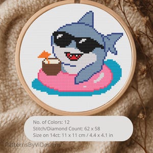 May include: A cross-stitch embroidery in a wooden hoop features a cartoon shark wearing sunglasses and floating on a pink inner tube. The shark is holding a coconut drink with an orange umbrella. The design is 4.4 x 4.1 inches.