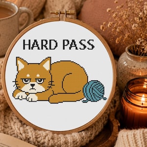 May include: A circular wooden embroidery hoop containing white fabric with a cross-stitch design of a grumpy orange cat lying next to a blue ball of yarn. The text HARD PASS is stitched in black capital letters above the cat.