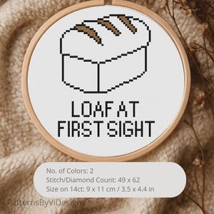 May include: A cross-stitch embroidery hoop featuring a pixelated loaf of bread design in brown and black on a white background. The text "LOAF AT FIRST SIGHT" is below the bread. The hoop is 3.5 x 4.4 inches.