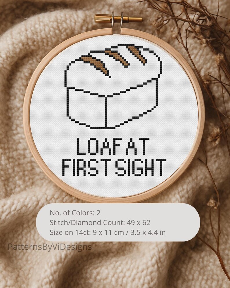 May include: A cross-stitch embroidery hoop featuring a pixelated loaf of bread design in brown and black on a white background. The text "LOAF AT FIRST SIGHT" is below the bread. The hoop is 3.5 x 4.4 inches.