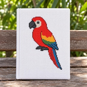 Può includere: Un taccuino rettangolare bianco con un ricamo a punto croce di un pappagallo ara sulla copertina. Il pappagallo è raffigurato con piume rosse, gialle, verdi e blu, un becco nero e una macchia bianca attorno all'occhio.