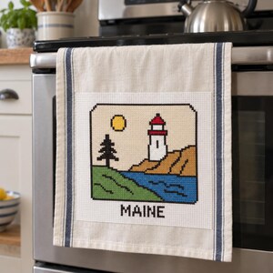 May include: A beige kitchen towel with dark blue vertical stripes featuring a cross-stitch design of a lighthouse, a pine tree, and a yellow sun over a blue body of water and brown land, with the word MAINE stitched in black at the bottom.