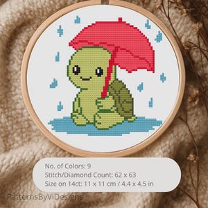 May include: A cross-stitch embroidery in a wooden hoop depicts a green turtle under a red umbrella, with blue raindrops. The design is 11 x 11 cm. The image text says "No. of Colors: 9" and "PatternsByViDesigns."