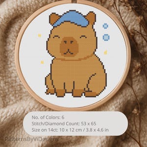 May include: A cross-stitch pattern in a wooden hoop featuring a brown capybara wearing a blue towel on its head, surrounded by small blue and yellow pixelated accents. The text includes No. of Colors: 6, Stitch/Diamond Count: 53 x 65, and Size on 14ct: 3.8 x 4.6 in.