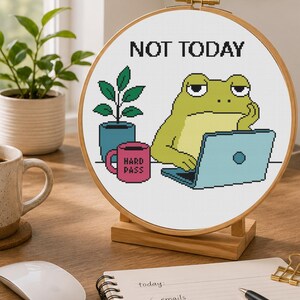 May include: A circular embroidery hoop containing a cross-stitch pattern of a green frog sitting at a light blue laptop. The text NOT TODAY is stitched above the frog, and a pink mug with the text HARD PASS sits next to a small potted plant.