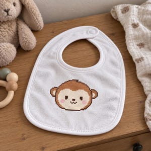 May include: A white baby bib featuring a cross-stitch style graphic of a brown monkey face with pink cheeks. The bib has a rounded shape with a snap closure at the top neck area and a white fabric border trim around the entire outer edge.